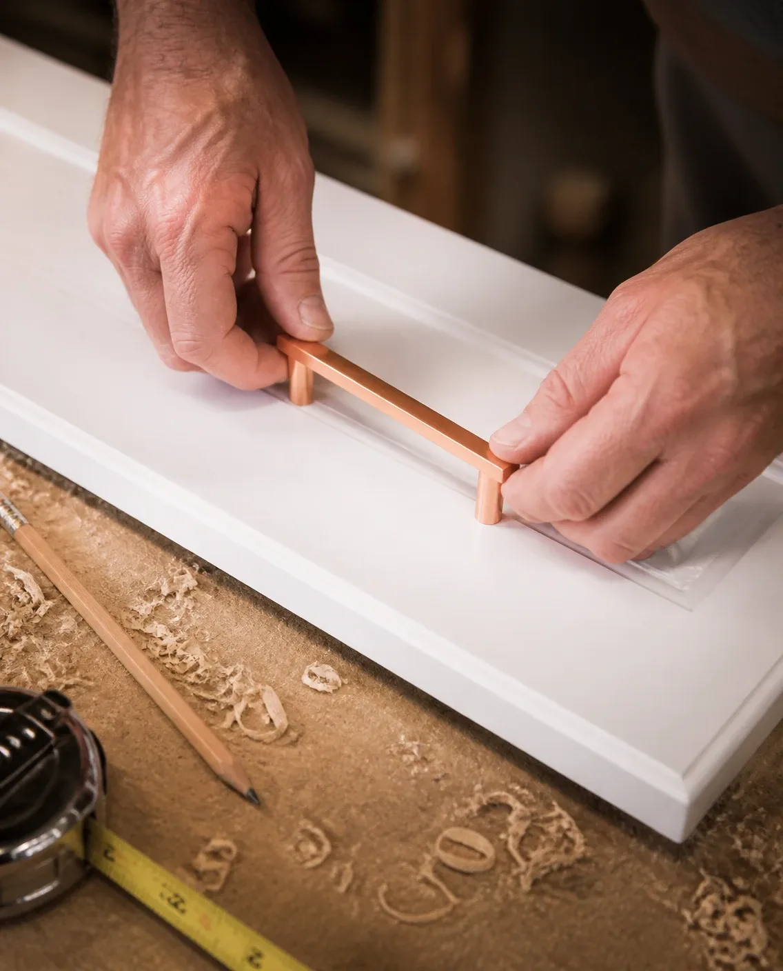 A craftsman's hands fitting a rose gold bar handle to a white cupboard door on a workbench
